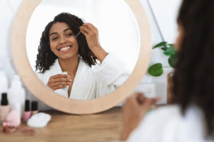 a beautiful black woman is applying a face serum in front of a mirror