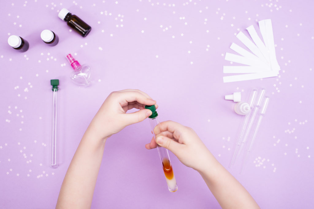 two hands holding a test tube with colorful liquid, small bottles of essential oils and a perfume bottle lying in the top corner, pipettes and test strips on the side